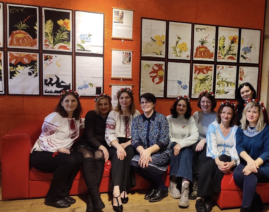 A group of Ukrainian women dressed in traditional clothing posing in front of the tapestries 